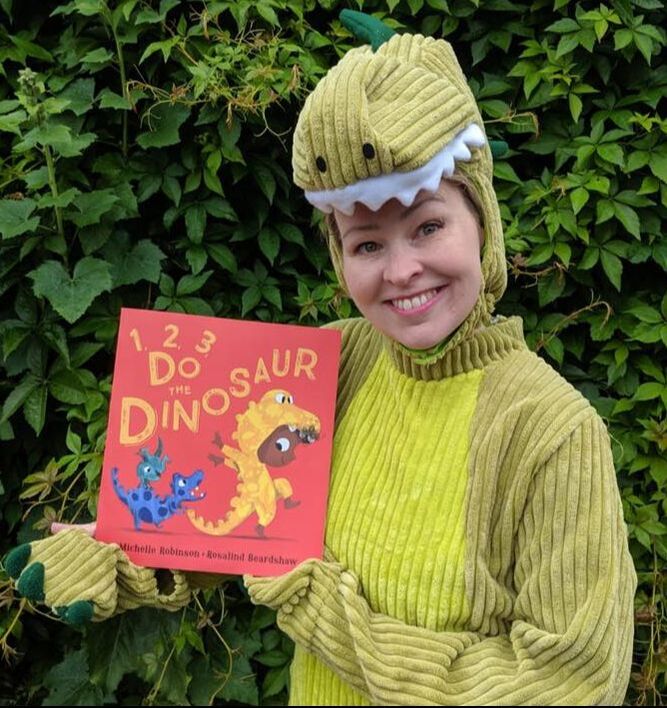 Author Michelle Robinson is dressed in a dinosaur onesie. She is holding her book 1,2,3 Do the Dinosaur and is smiling.