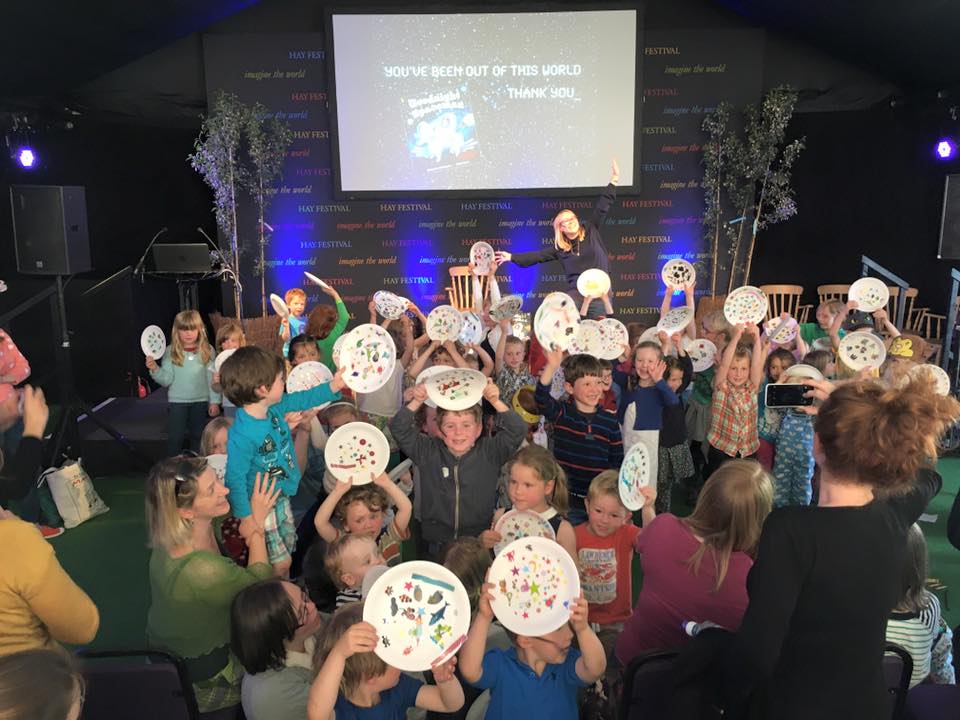 Michelle is performing in a marquee at Hay Festival. The room is packed with children, they hold aloft paper plates they have decorated as planets while Michelle spreads her arms wide.