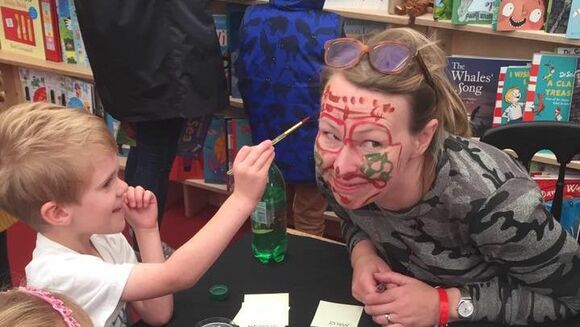Michelle is signing books in the Waterstones tent after performing at Cheltenham Literature Festival. She is leaning in to allow a mischievous little boy to apply face paint to her face with a paint brush.